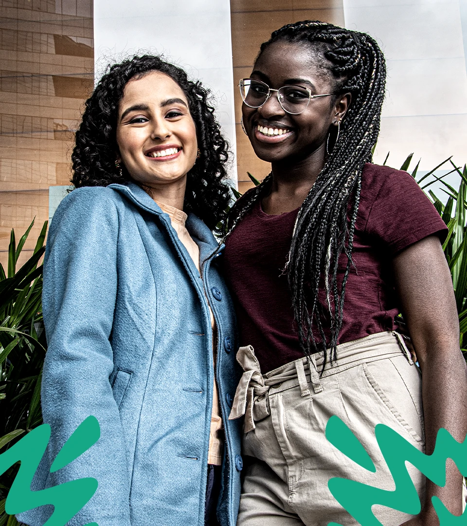 Foto de duas mulheres, esquerda uma mulher branca sorrindo com um blazer azul, mulher da direita, negra, blusa roxa escura e clara. Ambas estão sorrindo.