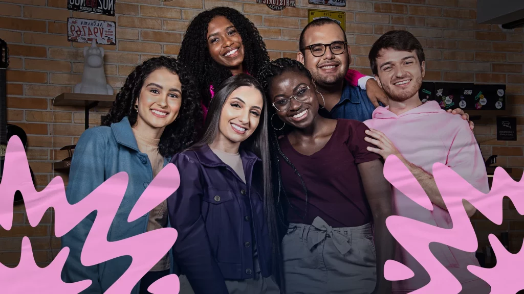 Foto posada de seis embaixadores do Programa de Estágio 2023. Da esquerda para a direita a foto é composta por uma jovem mulher parda com cabelo médio encaracolado, vestindo uma blusa creme com um casaco azul. Ao seu lado está uma mulher jovem branco com um cabelo longo e liso. Ela veste uma blusa branca com uma jaqueta roxa. Acima está uma mulher negra com cabelo longo encaracolado vestindo uma blusa rosa. Do lado está um homem pardo com cabelo castanho raspado e com barba rala, usando óculos e uma blusa azul jeans. Abaixo está uma mulher negra retinta, usando óculos e brincos de argola, com cabelos longos trançados. Ela usa uma blusa vinho e uma calça creme. Na ponta direita está um homem branco e loiro, com barba e vestindo uma blusa rosa clara. Todos estão sorrindo e atrás está uma parede de tijolos laranjas com quadros e decorações.